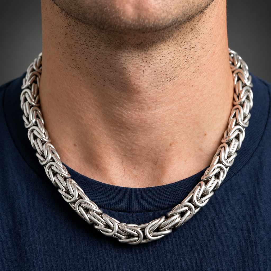 A close-up of a man's neck and chest wearing a thick, polished silver chain necklace with an intricate interlocking link design, paired with a dark navy blue t-shirt against a dark gray background.