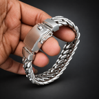 A close-up of a hand holding a silver-toned braided chain bracelet with a large rectangular clasp, set against a dark wood background.