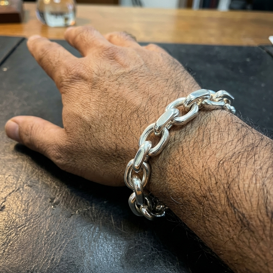 A close-up of a person's hand resting on a dark leather surface, wearing a chunky silver chain link bracelet.