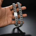 A close-up of a hand holding a thick silver Cuban link chain necklace with an ornate scrollwork pattern and a polished clasp, set against a solid black background.
