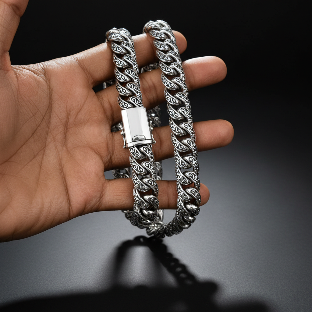 A close-up of a hand holding a thick silver Cuban link chain necklace with an ornate scrollwork pattern and a polished clasp, set against a solid black background.