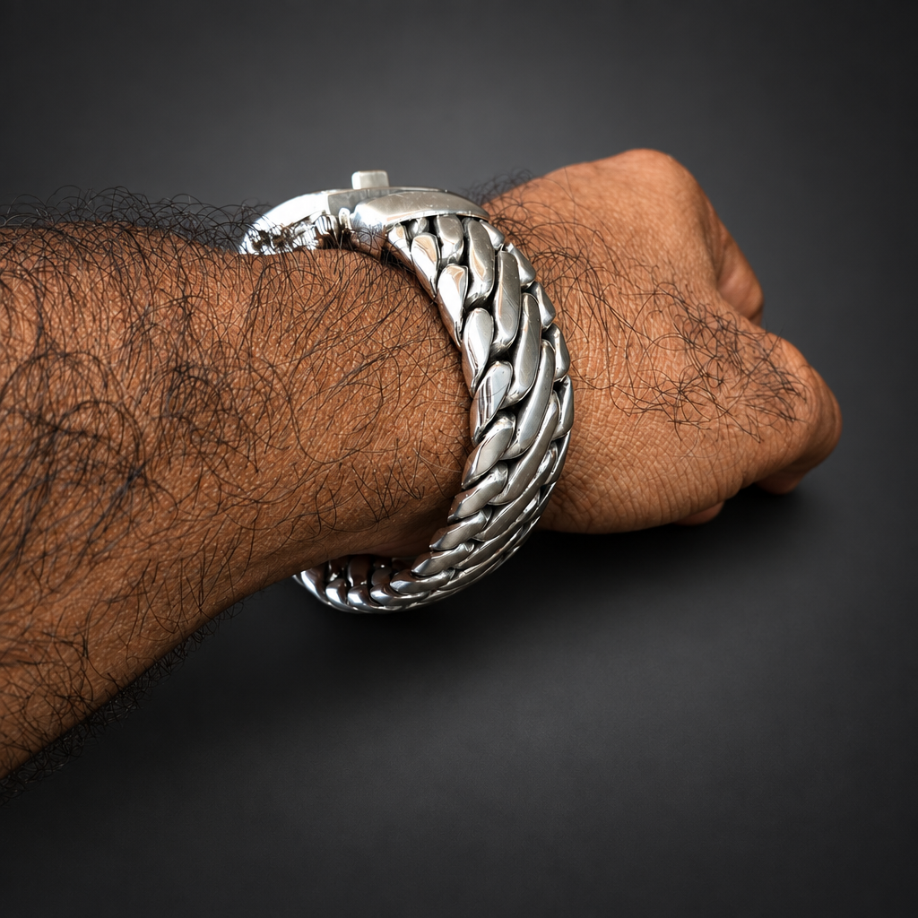 A close-up of a person's hairy forearm and hand wearing a chunky silver-toned braided chain bracelet, resting on a dark rustic wooden surface.