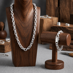 A silver chain necklace displayed on a dark wood bust alongside a matching silver bracelet on a smaller wooden stand, set on a gray fabric surface with blurred wooden jewelry boxes in the background.