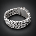 A chunky silver-toned metal bracelet with a woven curb-link design and a rectangular clasp, photographed on a dark gray to black gradient background.